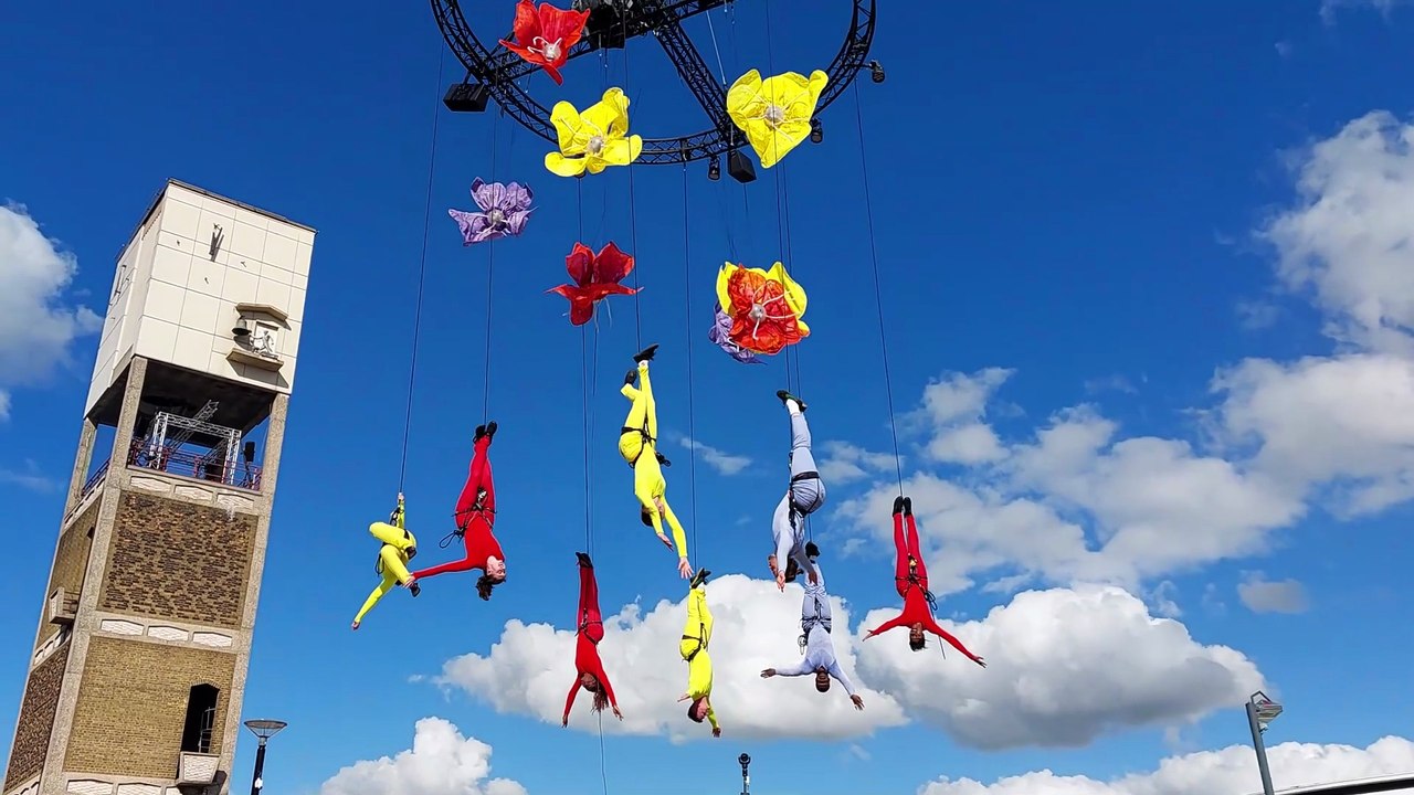 Brightly coloured people hanging from an arial structure against a blue sky background